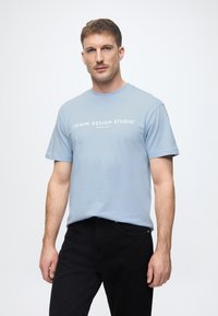 Ανοιχτό μπλε κοντομάνικο μπλουζάκι με στρογγυλή λαιμόκοψη, με λευκή εκτύπωση κειμένου. Συνδυάζεται με μαύρα τζιν και στέκεται μπροστά από ένα απλό φόντο.