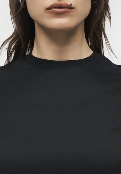 Κοντινό πλάνο του κάτω μέρους του προσώπου και του λαιμού μιας γυναίκας που φοράει μαύρο μπλουζάκι με στρογγυλή λαιμόκοψη, με ένα εμφανές ελιά πάνω από το άνω χείλος της.