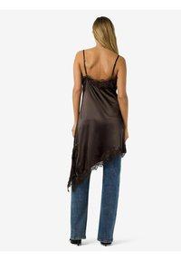 Sieviete ar gariem blondiem matiem, ģērbusies melnā asimetriskā satīna krekliņā ar mežģīņu apdari, valkā zilas džinsa bikses un melnus augstpapēžu apavus, skatās prom.