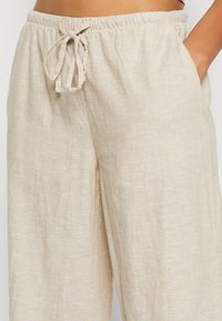 Beige linnebyxor med elastisk midja och knytband, med hand i sidofickan, bärs av en person mot en vit bakgrund.