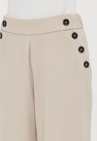 Μπεζ παντελόνι με εφαρμογή, κατασκευασμένο από απαλό ύφασμα, με ψηλή μέση και μαύρα κουμπιά στο πλάι για επιπλέον λεπτομέρεια.