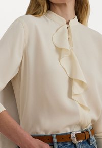Cream blouse with a ruffled placket and high collar, made of smooth fabric. Features pearl buttons and is paired with a brown belt.