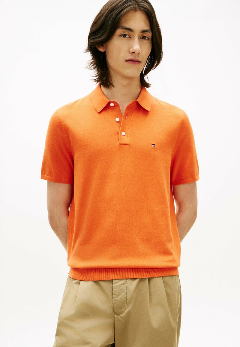 Jeune homme portant un polo orange à manches courtes et un pantalon beige, debout devant un fond blanc uni.