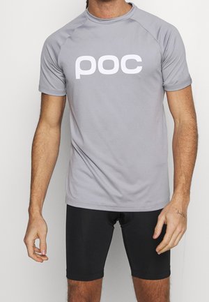 Homme portant un t-shirt gris clair à manches courtes avec le logo "poc" et un short de cyclisme noir sur un fond blanc uni.