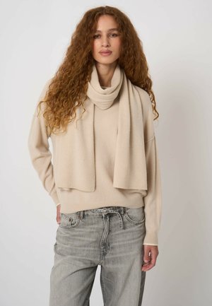 Kobieta o długich kręconych włosach, ubraną w beżowy sweter i pasujący do niego szalik, w komplecie z wysokim stanem jasnoszarych dżinsów, stojąca na tle jednolitego tła.