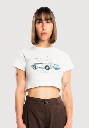 Giovane donna con capelli castani lisci che indossa un top corto bianco con un disegno di un'auto decappottabile blu e pantaloni marroni a vita alta.