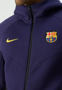 Paarse Nike zip-up jas met een hoge kraag, voorzien van een gouden swoosh-logo en een kleurrijk FC Barcelona-embleem op de linkerborst.