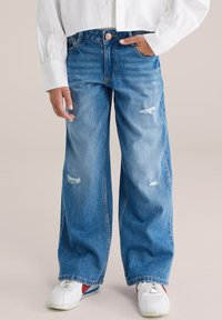 Persoană care poartă blugi largi albaștri cu mici rupturi, o cămașă albă cu mâneci lungi și adidași albi cu accente roșii și albastre.