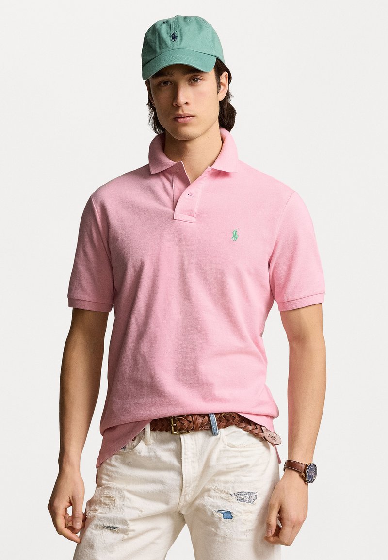 Camisa polo rosa clara de mangas cortas con un logotipo bordado en verde. Combinada con jeans blancos desgastados y un cinturón trenzado marrón. Gorra verde en la cabeza.