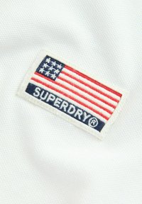 Vitt tyg med en texturerad yta har en broderad rektangulär lapp. Lappen visar den amerikanska flaggan och "SUPERDRY" i marinblått.