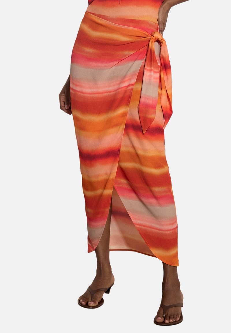 Femme portant une jupe portefeuille rayée orange vif, rose et beige avec un lien sur le côté, associée à des sandales marron à talons.