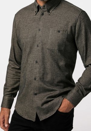 Man wearing long-sleeve, button-up gray herringbone shirt with chest pocket, one hand in black pants pocket, against plain background.