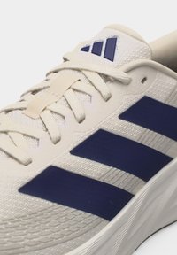 Макро снимка на бял спортен обувка с тъмносини ивици Adidas и връзки, завързани на панделка върху текстурирана горна част от мрежа.