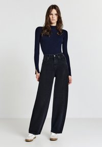Jeans neri a vita alta e gamba larga con tasche frontali, abbinati a un top a maniche lunghe aderente a righe blu navy e sneakers bianche su uno sfondo neutro.