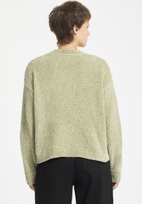 Pull en tricot vert clair avec une finition texturée, col côtelé et épaules tombantes, associé à un jean noir.