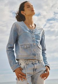 Lichtblauwe denimjas met een verkort ontwerp, metalen knopen, twee voorzakken en een ronde halslijn. Bijpassende lichtblauwe denim hieronder voorgesteld.