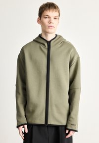 Mikina s kapucňou v olivovej zelenej l�átke, vybavená čiernym zipsom a manžetami, s jemným detailom loga. Voľný strih a mäkká textúra.