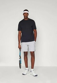 T-shirt preta de mangas curtas, calções cinzentos claros, faixa na cabeça branca, segurando uma raquete de ténis azul, a usar ténis brancos com detalhes azuis.