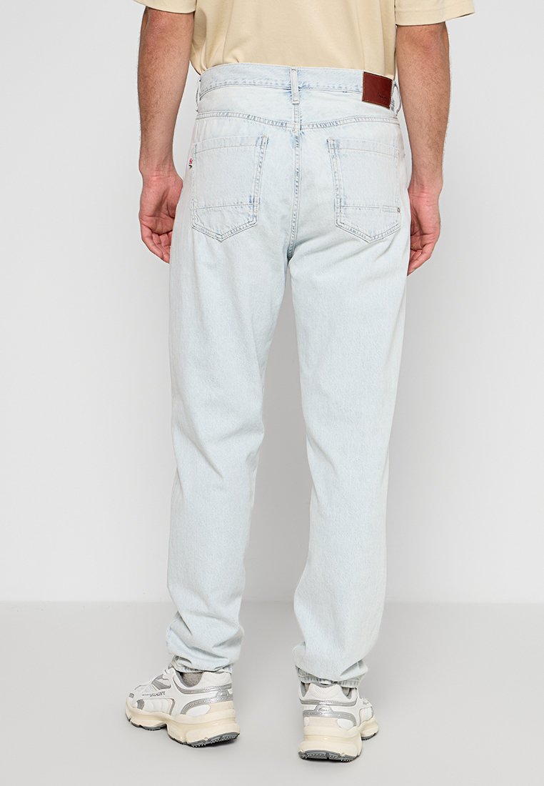 Hellblaue Denim-Jeans in lockerer Passform, mit Gesäßtaschen und einem braunen Lederpatch an der Taille. Getragen mit grauen Sneakers.