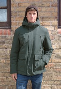 Dunkelgrüne isolierte Jacke mit hohem Kragen, zwei Fronttaschen und Kapuze. Modell trägt eine braune Strickmütze vor einer Backsteinwand.