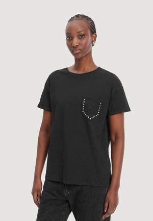 Jeune femme aux cheveux tressés portant un t-shirt noir avec une poche cloutée monogrammée "LV" et un pantalon foncé, debout devant un fond clair.
