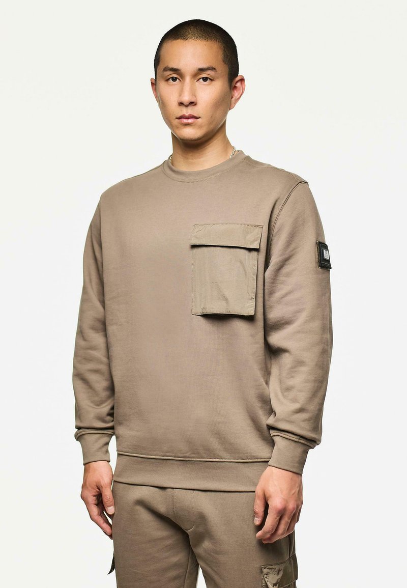 Uomo con felpa beige con grande tasca frontale e pantaloni abbinati, in piedi contro uno sfondo chiaro e semplice.