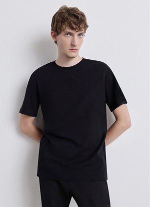 Junger Mann mit lockigem Haar, der ein schlichtes schwarzes kurzärmeliges T-Shirt und schwarze Hosen trägt, steht vor einer hellgrauen Wand.