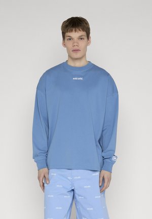 Junger Mann trägt einen lockeren blauen Sweatshirt und passende blaue Shorts mit wiederholtem weißem Logo-Schriftzug auf einfarbigem Hintergrund.