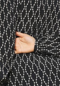 Sort stof med et flödende mønster prydes af hvide bølgede linjer og prikker. Ærmet er samlet ved manchetten, hvilket viser en glat tekstur.