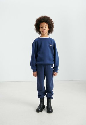 Tmavomodrá mikina a tepláky z mäkkej tkaniny, s logom na prednej strane a rebrovanými manžetami, doplnené čiernymi čižmami na zips.