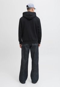 Uomo di spalle che indossa un cappellino azzurro chiaro, una felpa nera con il cappuccio abbassato e jeans scuri larghi, davanti a uno sfondo chiaro.