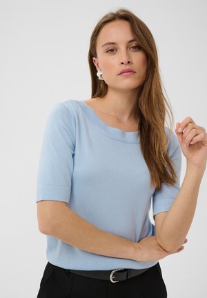 Femme aux longs cheveux bruns portant un haut bleu clair à manches courtes, des boucles d’oreilles argentées et un pantalon noir avec ceinture, posant sur un fond blanc uni.