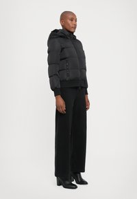 Femme debout dans une doudoune noire avec capuchon, pantalon noir à jambes larges et bottines à talons noires sur un fond blanc uni.