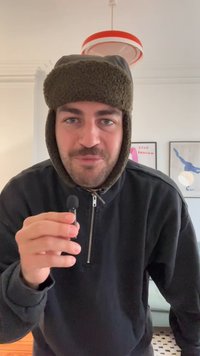 Fekete cipzáros dzseki, zöld szőrmebéléses sapka, egy kis fekete mikrofont tart. Egyszerű háttér, keretezett nyomatok a falon.