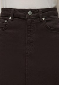 Jupe en jean marron foncé avec bouton sur le devant et passants de ceinture, portée avec un haut beige clair rentré.