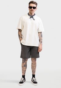 Άνδρας με τατουάζ στα χέρια και τα πόδια, φοράει γυαλιά ηλίου, λευκό φαρδύ μπλουζάκι, σκούρα σορτς, μαύρες κάλτσες, αθλητικά παπούτσια και μαντήλι γύρω από το λαιμό του.