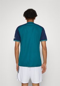Maglia atletica color teal con accenti blu navy, maniche corte e strisce laterali. Il tessuto liscio e la vestibilità comoda creano un design confortevole.