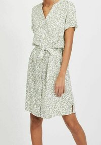 Femme portant une robe verte claire, à manches courtes et longueur genoux, avec des motifs abstraits blancs et une ceinture à la taille.