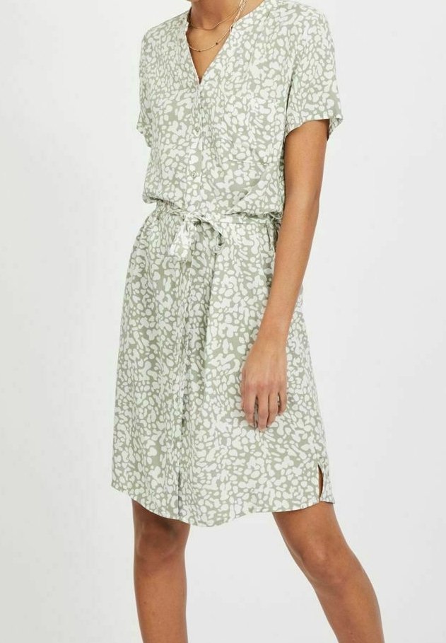 Femme portant une robe verte claire, à manches courtes et longueur genoux, avec des motifs abstraits blancs et une ceinture à la taille.