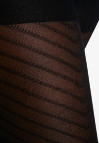 Meias pretas à riscas com um design transparente e texturizado. Apresentam padrões em zigue-zague tecidos no tecido, exibindo uma faixa preta lisa na parte superior.