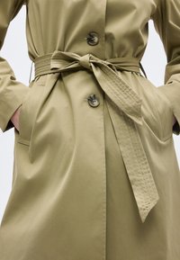 Γυναίκα που φοράει καφέ καμπαρντίνα με ζώνη δεμένη στη μέση, κλειστή με κουμπιά μπροστά, με τα χέρια στις πλαϊνές τσέπες μπροστά σε απλό φόντο.