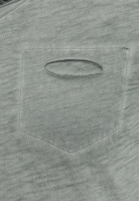 Jasnoszara bawełniana koszula z małą, owalną kieszenią z ukrytą nitką i teksturowanym wykończeniem. Tkanina ma subtelny wzór melanżowy.