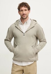 Felpa con cappuccio di un beige chiaro, caratterizzata da una tasca frontale, chiusura con zip e polsini a coste. Indossata sopra una maglietta a righe bianche e grigie.