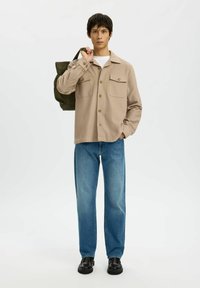 Jonge man staat, draagt een beige button-up shirt, blauwe spijkerbroek, zwarte schoenen en houdt een donkergroene tote bag over de schouder tegen een witte achtergrond.
