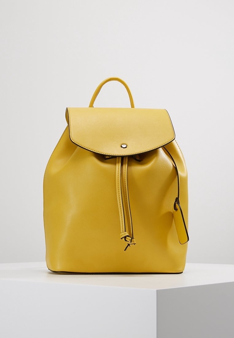 Sac à dos en cuir jaune avec un rabat supérieur, une fermeture à cordon et un bouton décoratif à l'avant. Texture lisse, forme arrondie et poignée unique sur le dessus.