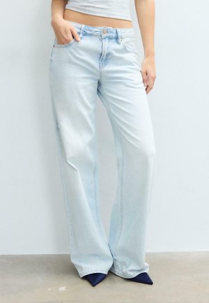 Femme portant un jean large bleu clair et un top blanc court, debout avec une main dans la poche, sur un fond uni.