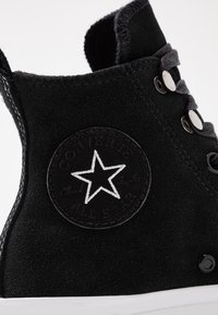 Schwarze Wildleder High-Top Sneaker mit einem kreisförmigen schwarzen Patch, einem weißen Stern und dem Text "Converse All Star", silbernen Ösen und weißer Sohle.