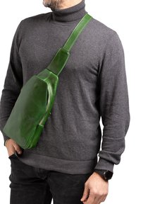 Borsa a tracolla verde in pelle con design piatto, dotata di chiusura a zip e una cintura ampia. La superficie ha una consistenza liscia.