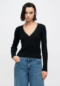 Μαύρο ριμπ πλεκτό ζακέτα με V-λαιμόκοψη και δαντέλα στο περίγραμμα, με κούμπωμα κουμπιών και εφαρμοστά μανίκια, σε συνδυασμό με ανοιχτόχρωμα τζιν παντελόνια φαρδιάς γραμμής.