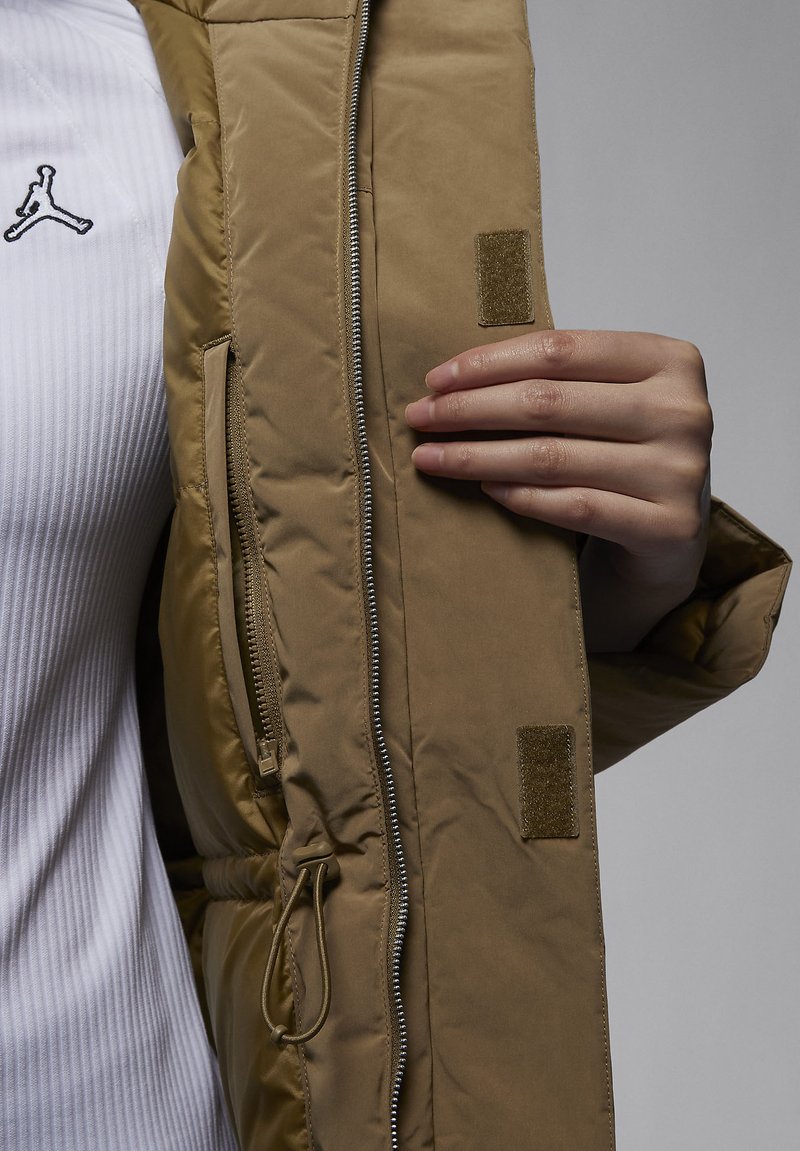 Chaqueta acolchada en caqui con una textura suave, cierre de cremallera y detalles de velcro. Cuenta con un bolsillo lateral y un cordón ajustable en la cintura.